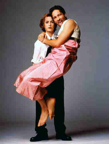photo of X-Files team Gillian Anderson and David Duchovny, she is dressed in heavy black boots, black pants and a white dress shirt with a black tie looking stern and lifting Duchovny off the ground, he is wearing a pink gown and sticking out his tongue