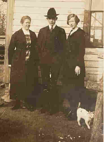 Black and white photo of three while people in dark clothing, posing together against the side of a light colored house. In the lower right corner of the photo is a petite shorthaired white and tabby cat. Its tail looks short like it may have been amputated at some point, but it seems healthy and happy.