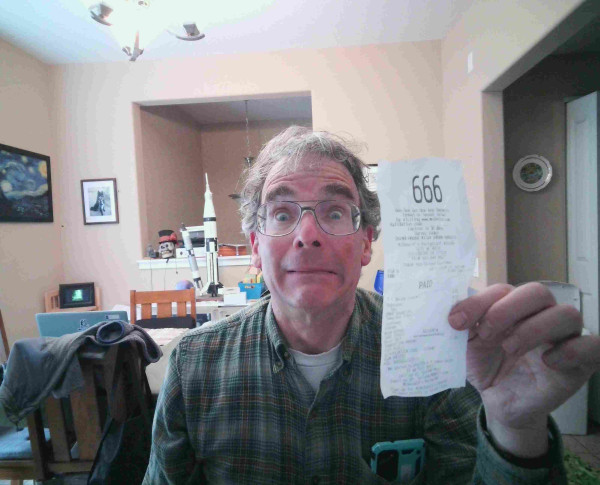 Middle aged man in a cluttered dining room, holding up a McDonald's receipt with the order number 666 printed on top.