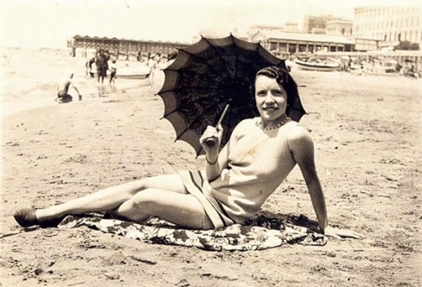 A black-and-white photo of a woman reclining on a sandy beach while holding an umbrella, with beachgoers and a pier in the background.
