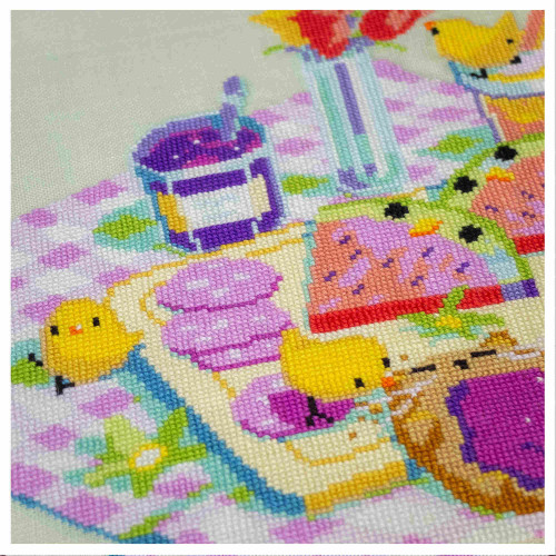 A zoomed in picture of the cross stitch picnic scene. There is a small chick taking some bites on a jelly toast. Another small chick is sitting on the napkin and looking in our direction.