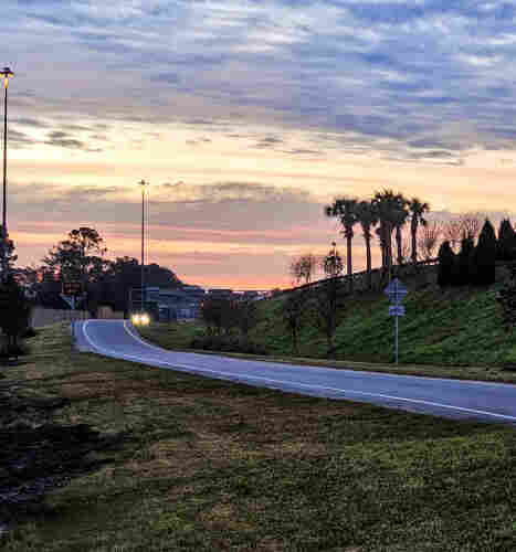 Looking out over the exit road for a highway pre-sunrise with little traffic, but those cars out have bright headlights on as they navigate the curved asphalt, with lush greenery to either side. Below a colorful sky with bands of purple, pink,  light yellow, bright white, and blue, and a thin blanket of grey-white clouds.