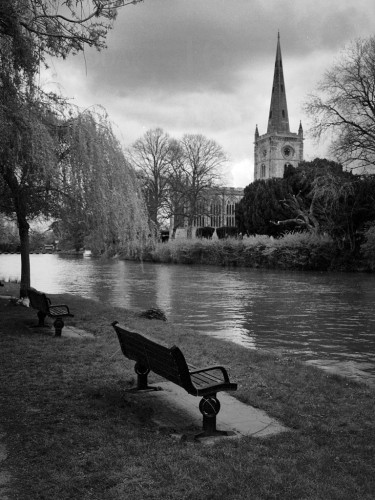 Black and white portrait format photo showing the tower and spire of the church at Stratford, across the Avon, under a moody sky. In the foreground is an ornate bench under a willow tree.