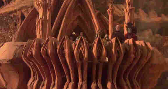 Still from Attack of the Clones depicting Count Dooku played by Christoper Lee and his alien allies standing upon a balcony which is decorated by architectural details which look like vulvas. Behind them is a vulva-shaped arch.
