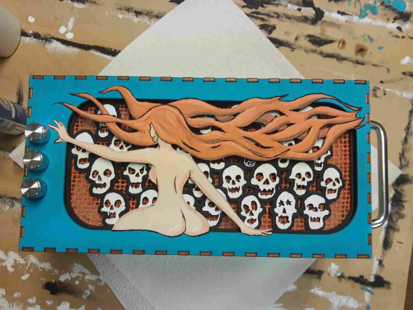Photo of lasercut speakerbox, with skulls and a pinup girl reaching for the power/ volume knob. Colors predominantly orange and teal