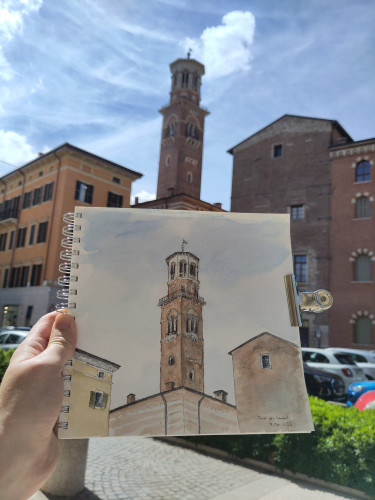 A ink&watercolor painting of Torre dei Lamberti in Verona in a sketchbook is held up by a hand. In the background is the real tower.
