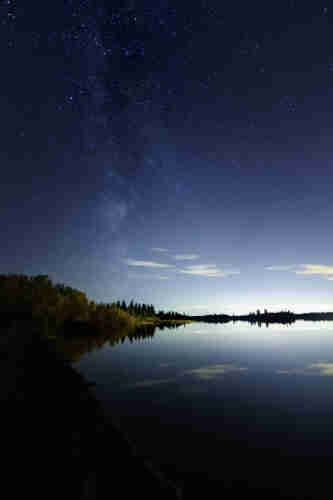 The Milky Way rises through a star-strewn blue sky, from deep blue at the top down to the palest hue along the horizon, where distant cityglow burns.  Faint wisps of white cloud obscure the sky just above the horizon; to the left, forest slopes down to the right and along the horizon. Below, a glassy, still lake reflects back the sky and forest. 