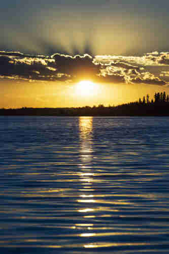 A glorious golden sunset sinks behind a line of grey-bellied white clouds, with rays stretching out above them into the blue sky. Below the clouds, a line of distant dark silhouetted forest separates a vivid and deep blue lake lined with ripples from the sky, through which the sun casts a long path of gold directly towards the observer. 