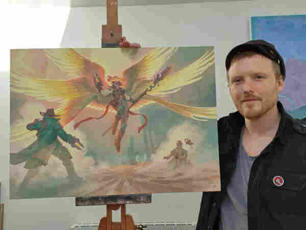 Angel of Indemnity painting with Denman Rooke posing on the right side. The angel floats above a dusty western street. 3 sets of wings sprawled out kicking dusty into the air. Two duelists hold their hands up to protect against the dust with one on the ground bleeding. The angel looks majestic with a halo around her face covering her eyes while adorning western attire and a wide brimmed cowboy hat.