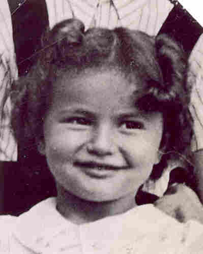 A photo of a face of a young smiling girl with dark hair pinned in the back and styled with few large curls. Someone is visible standing behind her.