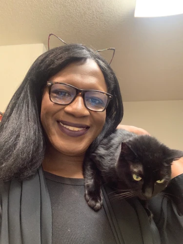 A dark-skinned woman with long black hair wearing wire-rimmed cat ears, abnd a black dress, holding a black cat. The black cat is on the woman's left shoulder.