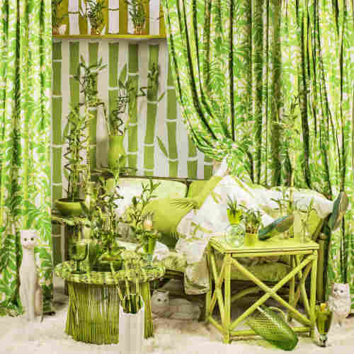 An elaborately staged photograph with a green theme featuring a posed mannequin disappearing into a couch and overwhelmed by a chaotic scene of bamboo-themed furnishings.