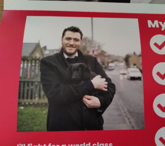 A bit of an election campaign leaflet. There is a picture of a smartly dressed white man with dark hair and a short beard standing by a road, printed on red paper. The man is holding a young black spaniel, I think probably a springer spaniel. The spaniel is adorable.