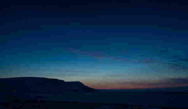 Sunset skies in Victor Bay. Low on the horizon the sky is a deep orange, lightening as you look higher. Then comes a light light blue that gradually deepens to a dark indigo. Barely visible at this resolution Jupiter shines above the mountains on the left. Scattered thin clouds are a deep pink. 