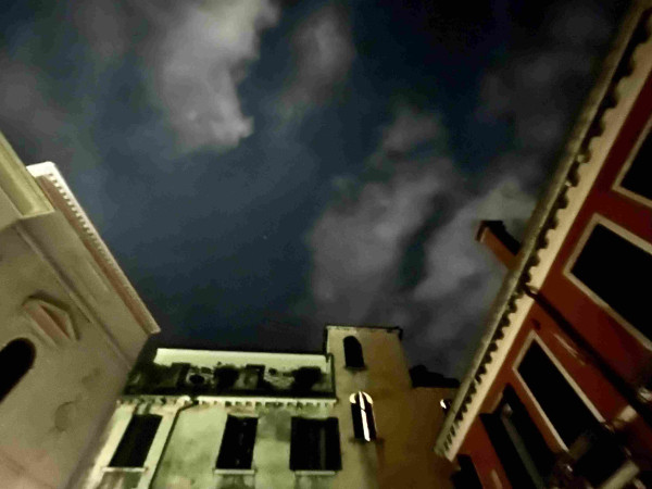 Colour photo taken late at night, looking up at the top storeys of three buildings which form three sides of a courtyard. The building on the left is white marble, and has one small arched dark window, the building on the right is painted scarlet, and has a row of long narrow rectangular shuttered windows, and the building straight in front of the camera is a slightly eerie creamy-green colour. The windows on this central building are all shuttered and dark apart from one, which is has its shutters partially open, and the warm light from within seen through this narrow slit and bouncing off the arch above makes the shape of a glowing arrow. The sky above is dramatically glowering dark blue with some white streaks of cloud. 