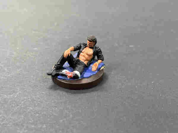 A painted miniature of Dr. Ian Malcolm from the original Jurassic Park movie, lounging on a seat cushion with his shirt fully unbuttoned after having been injured.