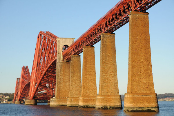 The Forth Bridge. The image shows the Forth Bridge seen from the south-west with evening sun illuminating its whole length. There are five stone supports at the near end, leading in from the right of the frame, and these are lit by the sun to a bright honey colour. The metal structure of the bridge is in the lower left of the frame and is bright reddish-orange. The sky is blue, and this is reflected in the water.