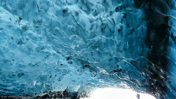 A photo taken from inside a cave. The low ceiling is comprised of thick, semi-transparent turquoise and blue ice. Dark areas can be seen which correspond with patches of snow on the other side of the ice. The surface of the ice is rippled, almost like the inverted surface of choppy water. The opening to the cave is at the bottom right of the shot, and a person can be seen walking in. All around the entrance point, light is being picked up by the ice roof above. There is another person standing at the bottom centre of the shot, only their helmet is visible.