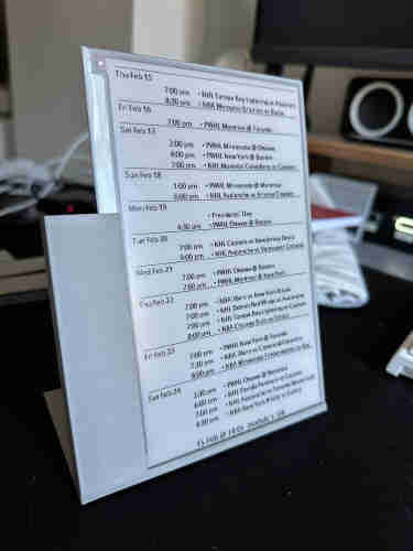 A bare epaper glass display with a list of upcoming sports games on it. It’s held at a 20% angle to the vertical with a grey plastic printed part that holds the display on the bottom and left sides