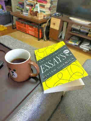 The yellow paperback book with squiggly black lines sits on the edge of a couch divider. To the left is a brown mug of black coffee with a LINE app brown bear mascot on it. The photo is in a living room