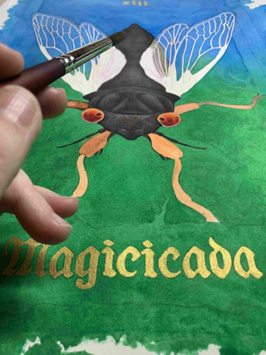 Painting in progress of a red eyed black cicada on a green and blue background. The genus name Magicicada is painted in gold and a hand holding a paintbrush is in the foreground. 