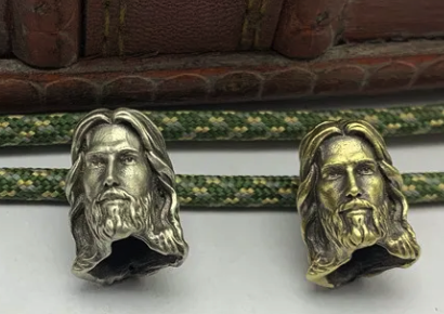 Two brass toned beads meant to represent Jesus. He is a bearded white American looking guy with long hair. 
