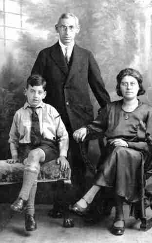 Studio family photo. On the left sits a boy in long socks, short trousers, with a tie and wearing braces. Behind him stands his father dressed in a suit. He has grey hair and glasses. On the right, a woman in a dark dress sits on a chair. She has dark hair. They are all looking straight into the camera.