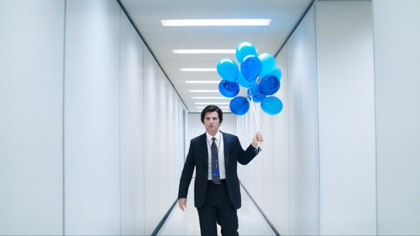 From Severance Season 2. Adam Scott as Mark S. walks the stark, vacuous white halls of Lumon Industries wearing his familiar black suit. He sports a flummoxed expression, in sharp contrast to the joyous bouquet of light and dark blue balloons he holds in his left hand. On his tie is a circular blue pin.