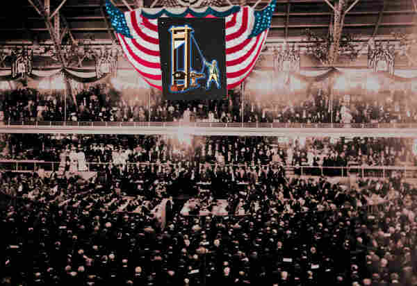 A black and white photo of a massive political party convention (the 1904 GOP convention); suspended over the stage, framed by US flag bunting, is a giant image of a French revolutionary operating a guillotine.