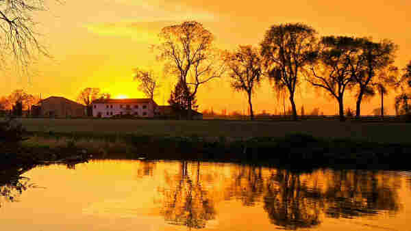 A serene sunset bathes the landscape in a warm, golden glow. The day's end brings a hush over the countryside, where a still pond mirrors the sky's fiery colors and the silhouette of leafless trees. A traditional farmhouse and outbuilding stand in quiet repose, their simple forms outlined against the luminous sky. This moment of tranquility is a silent symphony, celebrating the day's gentle closure.