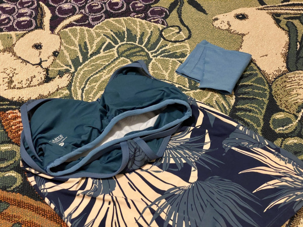 Horizontal photo of my tankini on the woven cotton bedspread with vegetable fruit and rabbit designs, showing the modified shelf bra bottom. The tankini is navy blue and has white and blue tropical plant designs. The folded blue legging material I used for this is next to it.