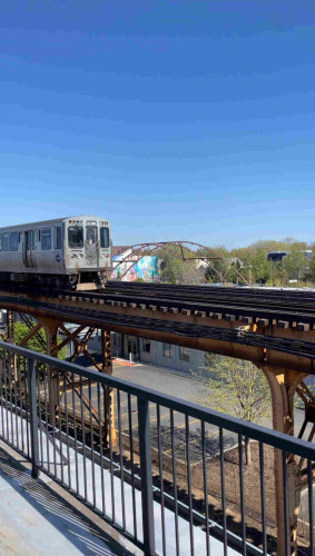 The Chicago el train running on a train very close to a black metal railing of the roof I’m on. In the background is a little bridge over a walking trail and a clear blue sky 