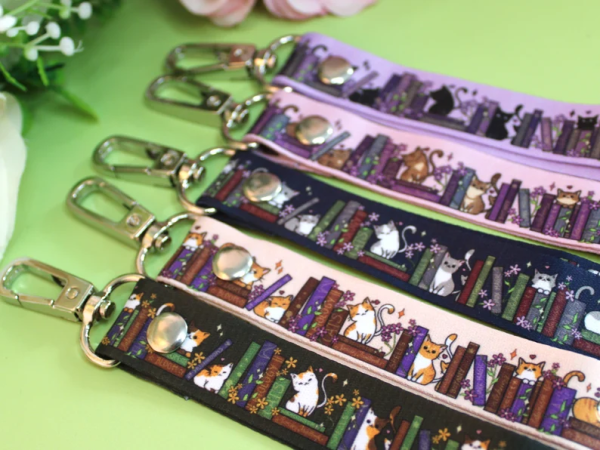 Lanyards with little cats sitting around and on top of books. Five colors: Top is purple and black cats, second is pink with brown cats, third is dark blue with silver cat, fourth is beige with orange cat, last is dark green with calico cats.