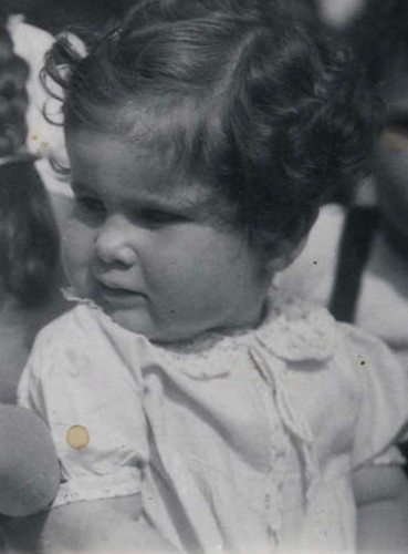 A vintage black and white photo of a young child with curly hair, wearing a delicate dress, focused intently on something out of frame.