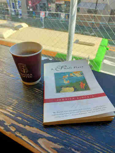 The photo is of a worn wooden counter against a second floor window. The white paperback book with an illustration of a painting of black children holding a white sheet to catch blond white girls in white nightgowns falling from a branch while a white male troubadour looks on from the right. To tye left is a brown paper cup of black coffee with Yanaka Coffee and its logo on it. Outside the window you can see shops across the street and the top of a bus directly below