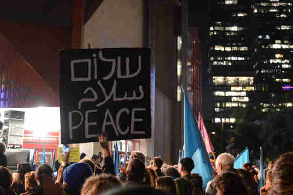 Crowd of people. A hand raised up holding a black sign that reads “Peace” in Hebrew, Arabic and English.