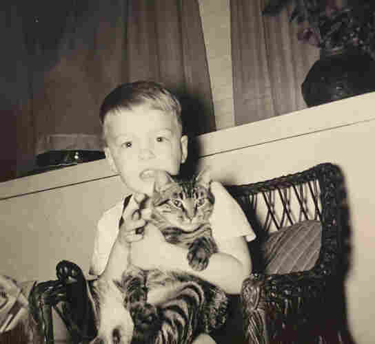 Black and white photo of a young white boy with blonde hair sitting in a wicker chair, holding a shorthaired tabby cat in a seated position in his lap. The cat seems fine with it, the body language is relaxed.