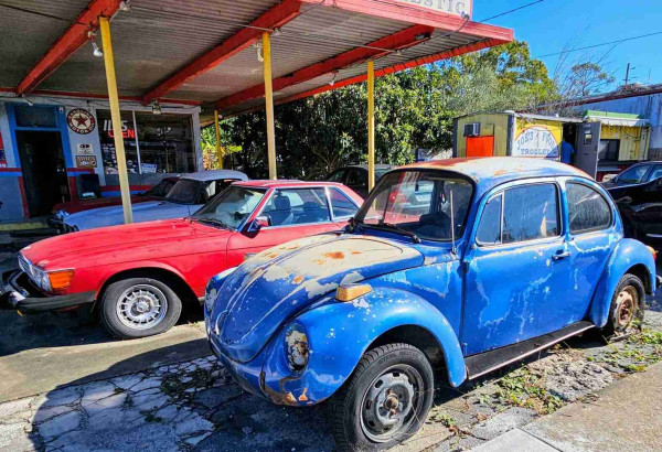 A "junk-yard" looking entrance for an auto repair shop, with numerous disabled vehicles. In the front a 1972 Volkswagen Beetle in blue, has seen better days but is still quite stunning.