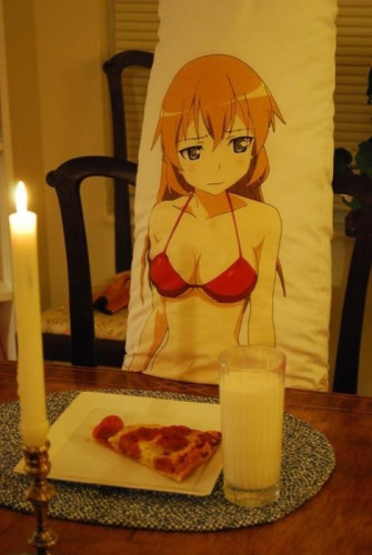 Waifu pillow sitting at the dinner table with a lit candle, a slice of pizza, and a glass of milk on the table