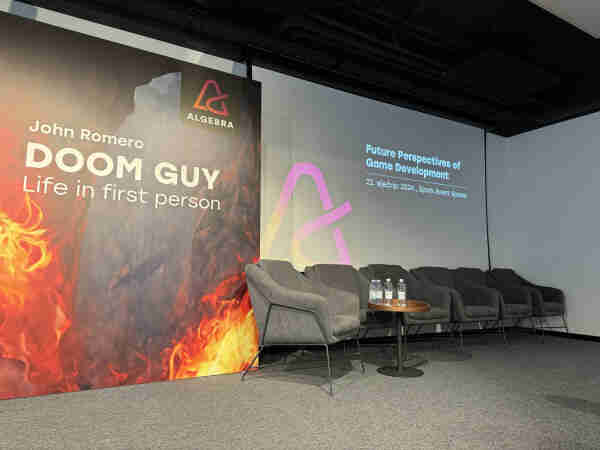 Event stage with chairs, a banner for John Romero, "DOOM GUY - Life in first person," and a screen with text "Future Perspectives of Game Development."