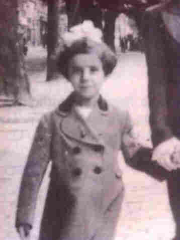 A young girl photographed in the street. She is wearing a coat and holding some adult's hand - probably her mother. She has white bow in her hair. The hair reach to her ears. Trees and park lane is visible in the background.