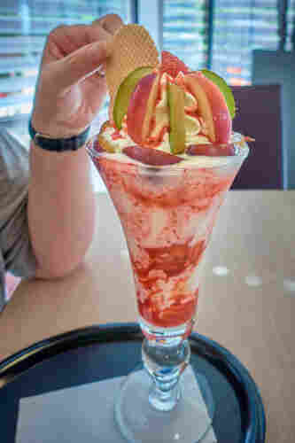 A large strawberry sundae sits on the table. The topping of whipped cream is decorated with kiwi and apple slices and a round waffle. One hand is grabbing the waffle...
