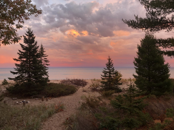 A serene beach scene with pine trees, dunes, and a calm lake under a colorful sunset sky.