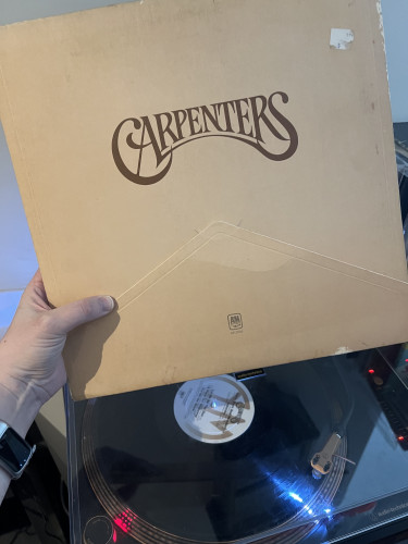 My hand holding the album cover to The Carpenters' 1971 album, also called Carpenters, above the record playing on a turntable 
