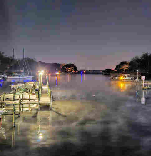 Late night overlooking a small marina at the mouth of Goodby's Creek and the Saint Johns River. Red decorative lights outline portions of the marina's shoreline features. Blue lights outline a nearby dock and a smaller one on the far shoreline across the water. Yellow lights appear at the rear of some boats on both sides of the water. Bright city lights can be seen far off in the distance beyond the creek, and the vast river the creek flows into. All brightly illuminated and reflecting upon the water below a blue night sky.