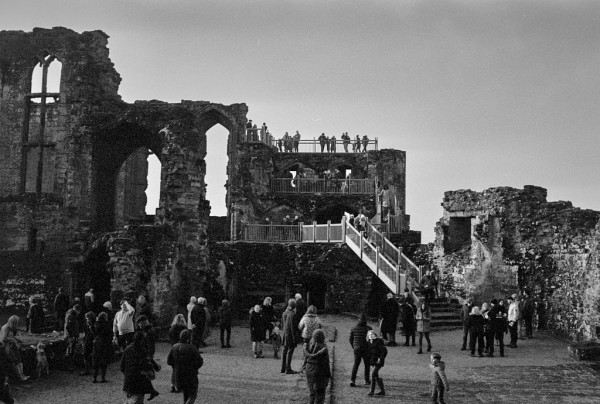 The walls of a high stone castle, from inside, with dozens of people milling around. A wooden stairway leads up to the top of one of the walls, where people stand to admire the view. Some of the walls have tall windows in them. Black and white photo.