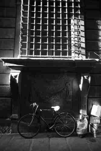 Bike against exterior wall, a window above covered by security bars, in black and white, capturing light and shade. Debris awaiting collection to the right.
