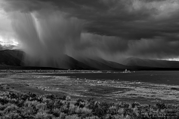 A black and white landscape photo of a rain squall over the foothills of a mountain range. In the foreground is sagebrush, a lake's shoreline, lake water to the right, a line of short spiky rock formations extending out into the lake. In the background are dark clouds pouring rain along a line of dark mountains. A bit of bright light is seen in the center left edge of the frame. 