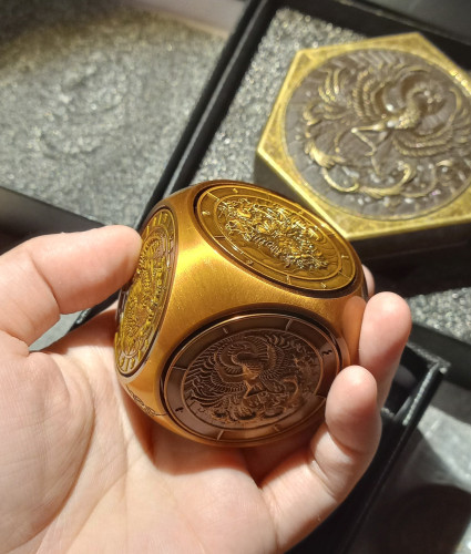 Hand holding a large rounded cube about the size of a nectarine. The cube has a shiny polished gold color finish. Each face is a circular dial with a different mythological creature on it and the rim of each dial is marked off in segments corresponding to the sides of a polyhedral die.