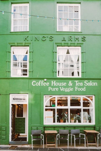 A green building fills the frame, with white wood-panelled windows. Letters across the top painted in green read 'King's Arms'. Further down at street level there's an open doorway, a bigger panelled window showing warm lighting inside, some tables and chairs out the front, and white painted text on the wall above the window reading 'Coffee House & Tea Salon, Pure Veggie Food'.